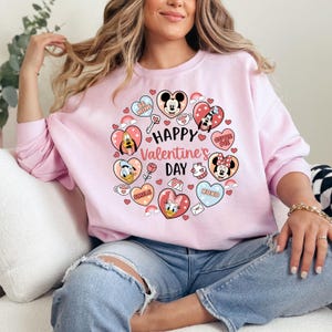 K&ouml;nnte beinhalten: Ein hellrosa Sweatshirt mit einem Valentinstagsdesign. Das Design zeigt einen herzf&ouml;rmigen Kranz mit Zeichentrickfiguren, Herzen und dem Text "Happy Valentine's Day". Das Sweatshirt hat lange &Auml;rmel.