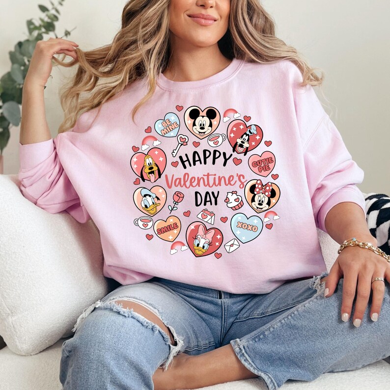 K&ouml;nnte beinhalten: Ein hellrosa Sweatshirt mit einem Valentinstagsdesign. Das Design zeigt einen herzf&ouml;rmigen Kranz mit Zeichentrickfiguren, Herzen und dem Text "Happy Valentine's Day". Das Sweatshirt hat lange &Auml;rmel.