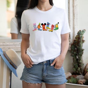 May include: White t-shirt featuring a colorful graphic of cartoon cowboy boots. The boots are in various colors and patterns, including pink, blue, red, and yellow. The shirt is paired with denim shorts.