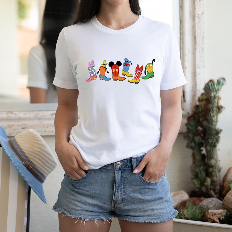 May include: White t-shirt featuring a colorful graphic of cartoon cowboy boots. The boots are in various colors and patterns, including pink, blue, red, and yellow. The shirt is paired with denim shorts.