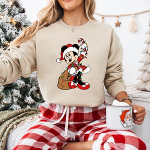 May include: Beige sweatshirt featuring a Christmas-themed Mickey Mouse graphic. Mickey is dressed in a Santa suit, holding a candy cane and a sack of gifts, surrounded by Christmas lights. The sweatshirt is paired with red and white plaid pajama pants.