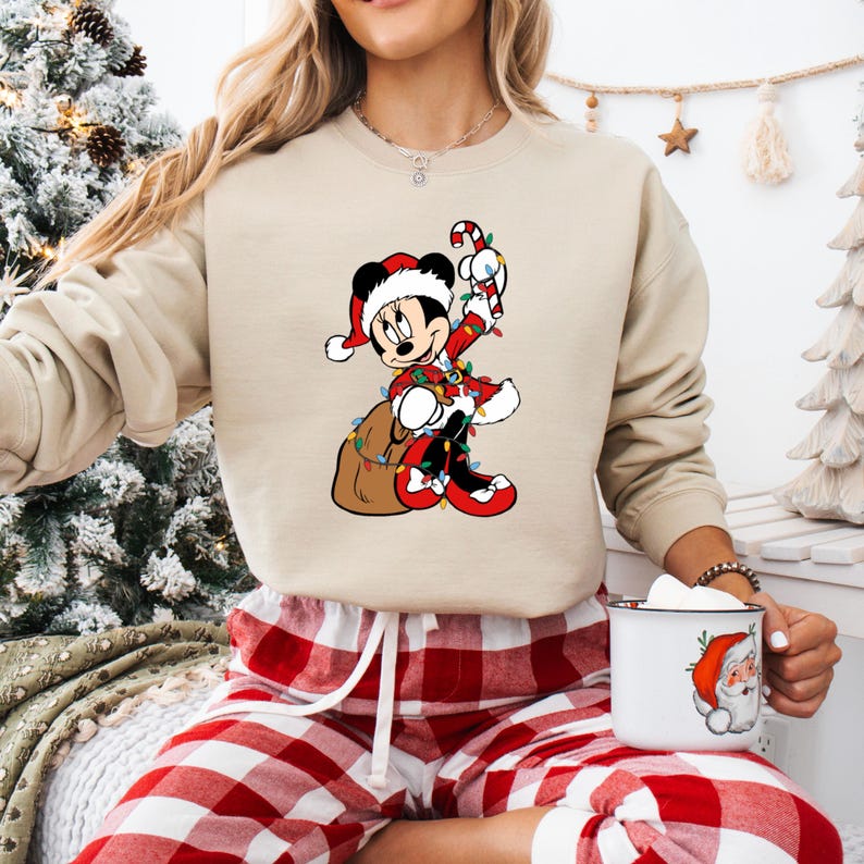 May include: Beige sweatshirt featuring a Christmas-themed Mickey Mouse graphic. Mickey is dressed in a Santa suit, holding a candy cane and a sack of gifts, surrounded by Christmas lights. The sweatshirt is paired with red and white plaid pajama pants.