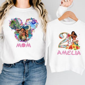 May include: White long-sleeve sweatshirts with matching designs. One features a Mickey Mouse head with a tropical scene and the word "MOM" in pink. The other has a number 2 with a character and the name "AMELIA".
