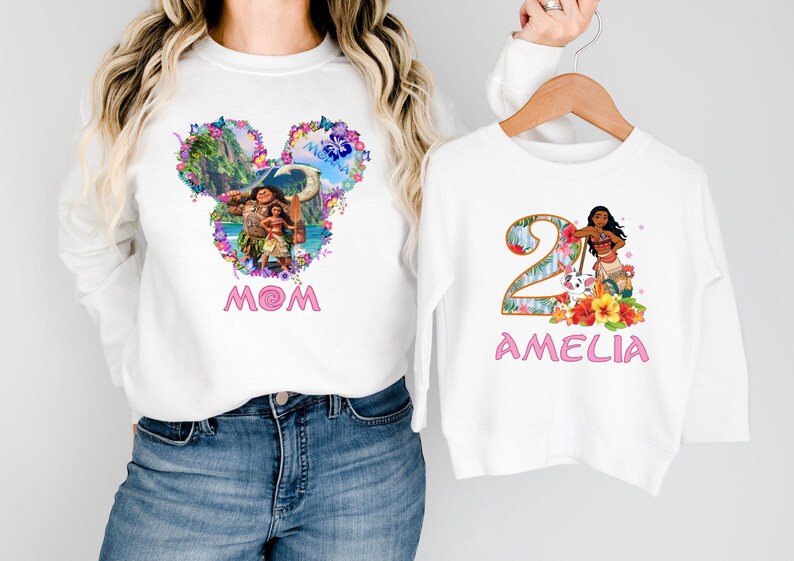 May include: White long-sleeve sweatshirts with matching designs. One features a Mickey Mouse head with a tropical scene and the word "MOM" in pink. The other has a number 2 with a character and the name "AMELIA".