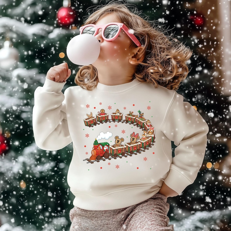 May include: Cream-colored sweatshirt featuring a festive train design with gingerbread men and cartoon characters. The train is red and green, with a white cloud of smoke. The design includes red and green snowflakes. The sweatshirt is perfect for the holiday season.