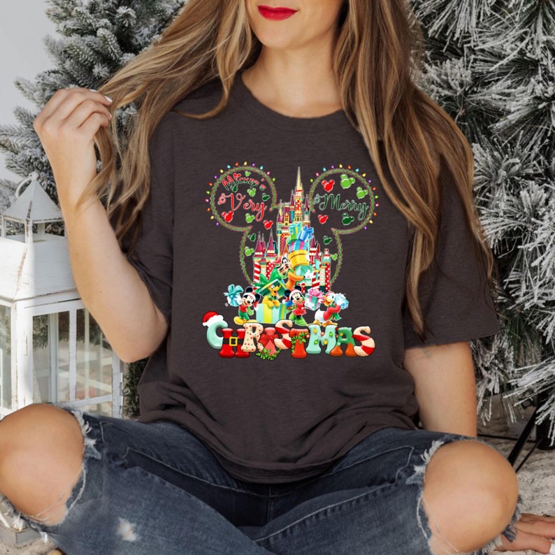 May include: A dark grey t-shirt with a Christmas design featuring Mickey Mouse ears, the Disney castle, and characters such as Mickey Mouse, Minnie Mouse, Donald Duck, Goofy, and Pluto. The text "Happy Very Merry Christmas" is written on the shirt.