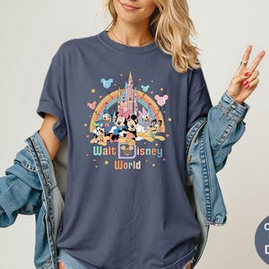 Puede incluir: Camiseta azul marino con un dise&ntilde;o gr&aacute;fico colorido de personajes de Disney frente a un castillo y un arco&iacute;ris. El texto "Walt Disney World" est&aacute; debajo. La camiseta es Comfort Colors Denim.