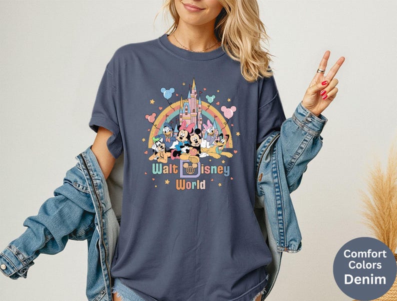 Puede incluir: Camiseta azul marino con un dise&ntilde;o gr&aacute;fico colorido de personajes de Disney frente a un castillo y un arco&iacute;ris. El texto "Walt Disney World" est&aacute; debajo. La camiseta es Comfort Colors Denim.
