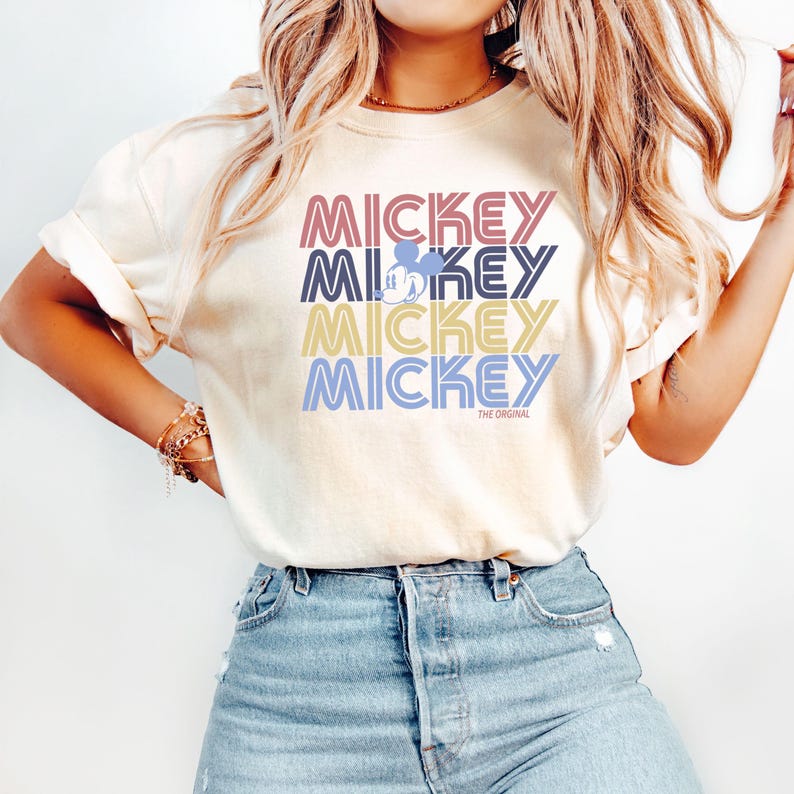 May include: Cream-colored t-shirt with the word "MICKEY" stacked four times in different colors: pink, blue, yellow, and purple. A small Mickey Mouse silhouette is in the second "MICKEY". The text "THE ORIGINAL" is below the last "MICKEY".