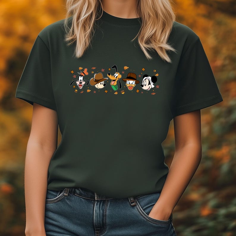 May include: Dark green t-shirt featuring a row of cartoon character faces, including Goofy, Mickey Mouse, and Donald Duck, each wearing a hat. The design includes autumn leaves. The shirt is worn by a person wearing blue jeans.