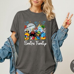 May include: Dark gray t-shirt featuring a colorful graphic with the numbers "2026" and Disney characters. The text "Smith Family" is below the graphic. The shirt has a crew neck and short sleeves.