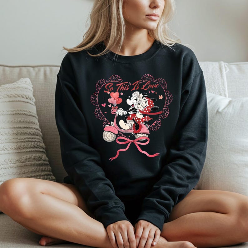 Puede incluir: Sudadera negra con un gr&aacute;fico de dibujos animados de una pareja en un scooter rosa, con el texto "So This Is Love" dentro de un dise&ntilde;o en forma de coraz&oacute;n. El gr&aacute;fico incluye corazones rosas y un gran lazo rosa.