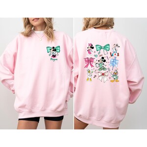 May include: A light pink sweatshirt featuring a Minnie Mouse and Mickey Mouse design with a teal bow. The back of the sweatshirt has multiple Disney character designs. The name "Megan" is printed below the front design.