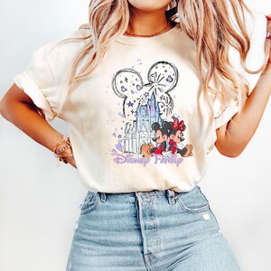 May include: Cream-coloured t-shirt with a graphic of Mickey and Minnie Mouse in front of a castle, with the words "Disney Family" in purple script. The design includes fireworks and stars. The shirt is worn with blue jeans.