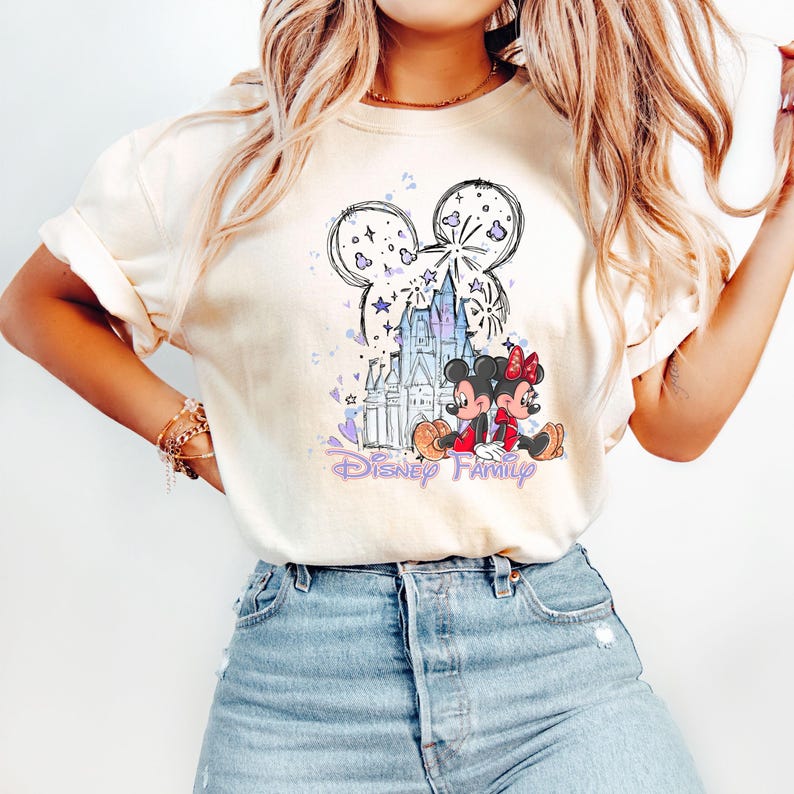 May include: Cream-coloured t-shirt with a graphic of Mickey and Minnie Mouse in front of a castle, with the words "Disney Family" in purple script. The design includes fireworks and stars. The shirt is worn with blue jeans.