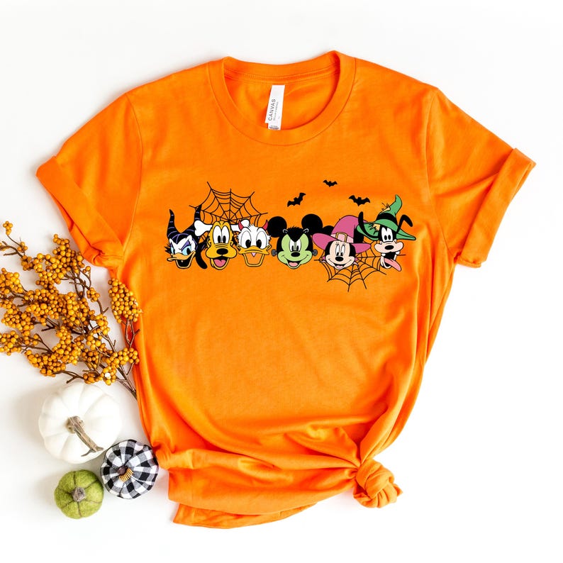 Pu&ograve; includere: Maglietta arancione con una grafica di Topolino, Minnie Mouse, Paperino, Pippo e Pluto vestiti con costumi di Halloween. I personaggi sono davanti a una ragnatela con pipistrelli che volano sopra di loro.