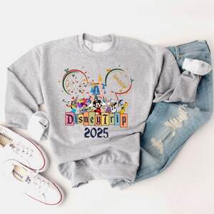 May include: A gray sweatshirt with a colorful graphic of Mickey Mouse and other Disney characters in front of Cinderella's Castle. The graphic also includes the text "Disney Trip 2025".