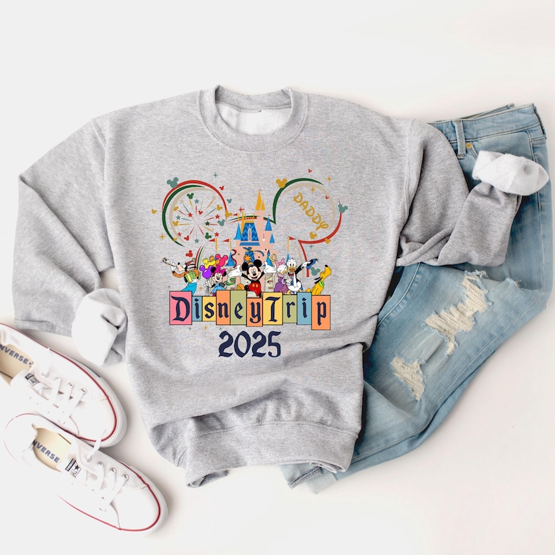 May include: A gray sweatshirt with a colorful graphic of Mickey Mouse and other Disney characters in front of Cinderella's Castle. The graphic also includes the text "Disney Trip 2025".