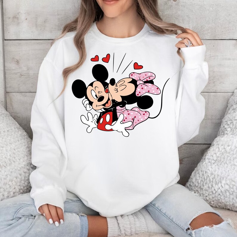 May include: White sweatshirt featuring a graphic of Mickey Mouse and Minnie Mouse kissing. The graphic is surrounded by red hearts.