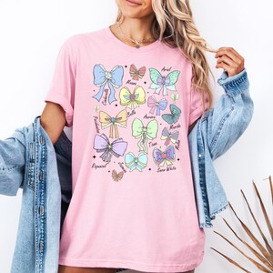 May include: Pink t-shirt with a graphic of bows and the names of Disney princesses: Ariel,  Mulan, Cinderella, Belle, Aurora, Merida,  Mulan, Tiana,  Jasmine, Snow White, Rapunzel.