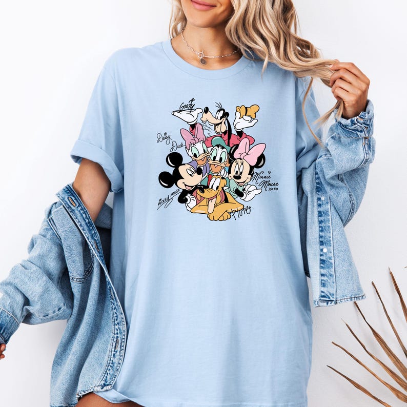 May include: Light blue t-shirt featuring a graphic of Disney characters including Mickey Mouse, Minnie Mouse, Donald Duck, and Goofy. The characters are surrounded by their signatures. The shirt is a light blue color.