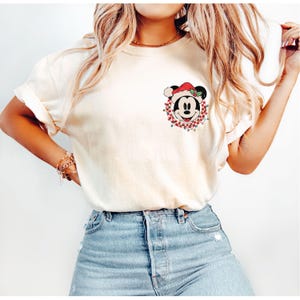May include: A white t-shirt with a Christmas-themed Mickey Mouse graphic. Mickey is wearing a Santa hat and is surrounded by a red and green wreath with lights.