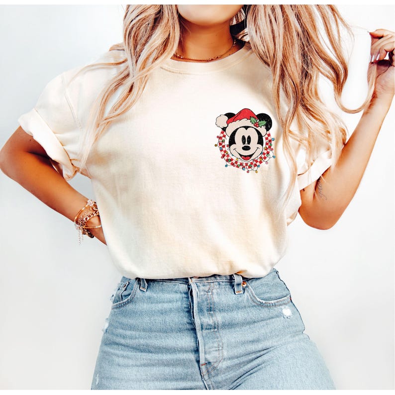 May include: A white t-shirt with a Christmas-themed Mickey Mouse graphic. Mickey is wearing a Santa hat and is surrounded by a red and green wreath with lights.
