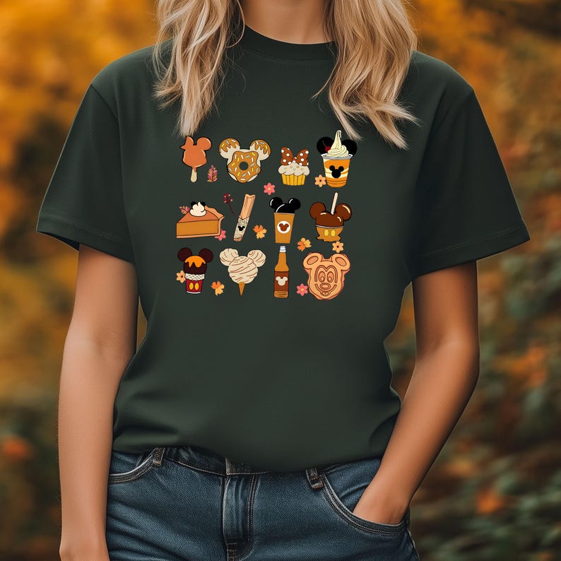 May include: Dark green t-shirt featuring a collection of cartoon food items with Mickey Mouse ears. The illustrations include ice cream, donuts, cupcakes, coffee, and cookies. The shirt is a solid color with a crew neck.