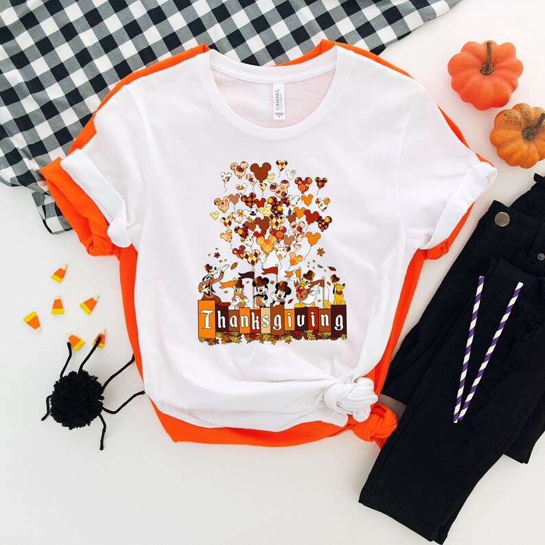 May include: White t-shirt with orange trim featuring a Thanksgiving design. The graphic includes Mickey Mouse-shaped balloons in fall colors, characters, and the word "Thanksgiving" in a decorative font. Black pants and pumpkins are also in the image.