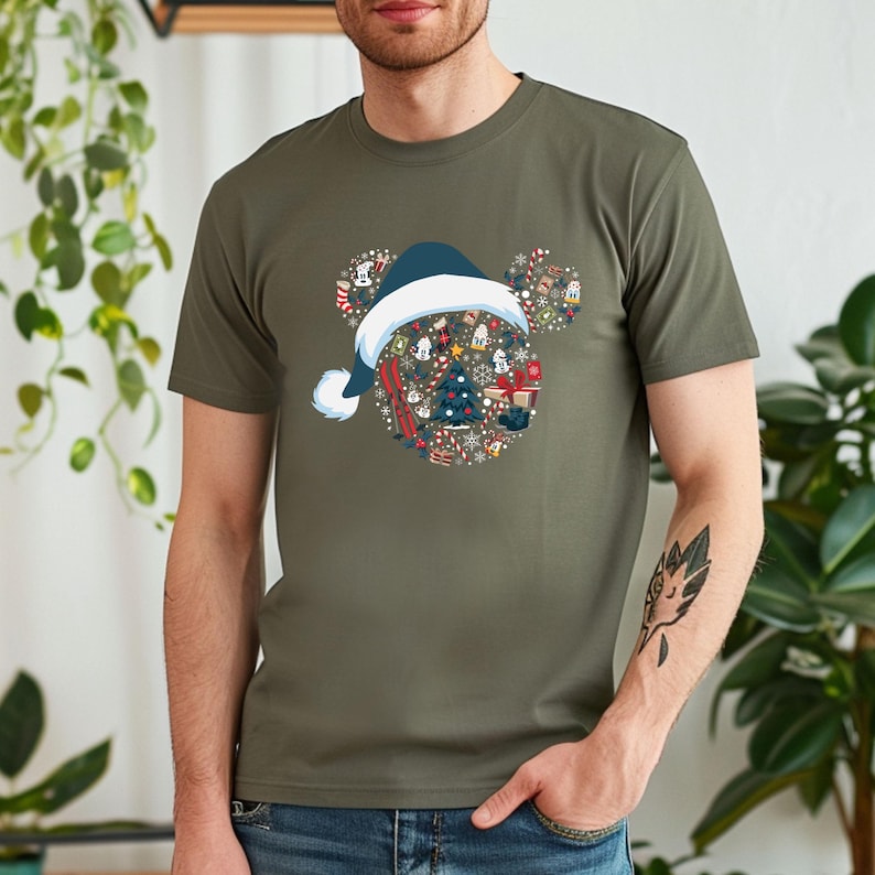 May include: Olive green t-shirt featuring a festive design. The design is a Mickey Mouse head shape filled with Christmas-themed elements, including a Christmas tree, skis, and snowflakes. A blue Santa hat with a white brim tops the design.