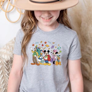 May include: A gray t-shirt featuring a colorful graphic of Disney characters, including Mickey Mouse, Minnie Mouse, Donald Duck, Goofy, and Pluto, with school supplies and autumn leaves. The shirt is likely for children.