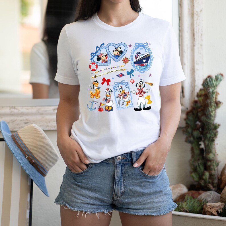 May include: White t-shirt with a colorful print of Disney characters, including Mickey Mouse, Minnie Mouse, Donald Duck, Goofy, and a cruise ship. The print features nautical themes like anchors, life preservers, and a ship's wheel.