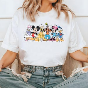 Puede incluir: Camiseta blanca con un estampado de dibujos animados de colores de Mickey Mouse, Minnie Mouse, Donald Duck, Goofy y Pluto. Los personajes est&aacute;n de pie en fila y sonriendo.