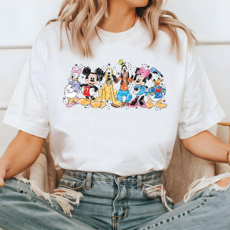 Puede incluir: Camiseta blanca con un estampado de dibujos animados de colores de Mickey Mouse, Minnie Mouse, Donald Duck, Goofy y Pluto. Los personajes est&aacute;n de pie en fila y sonriendo.