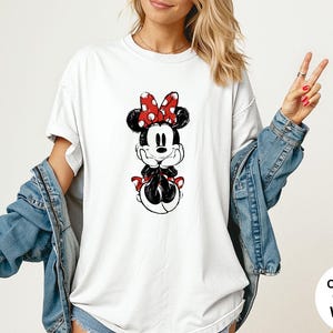 May include: White t-shirt featuring a black and red Minnie Mouse graphic. The design includes Minnie's iconic polka dot bow and outfit. The shirt is part of the Comfort Colors collection.