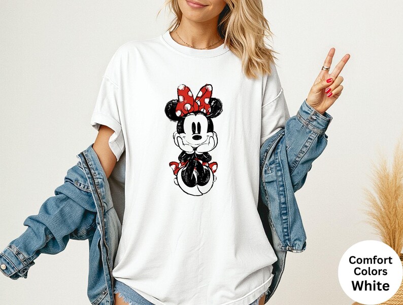 May include: White t-shirt featuring a black and red Minnie Mouse graphic. The design includes Minnie's iconic polka dot bow and outfit. The shirt is part of the Comfort Colors collection.