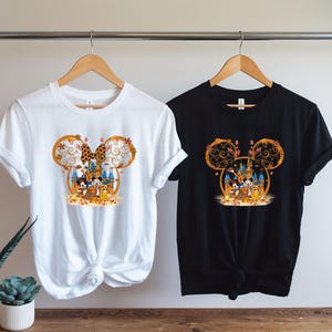 Pode incluir: Duas camisetas, uma branca e outra preta, cada uma com um design tem&aacute;tico da Disney. O design inclui um la&ccedil;o da Minnie Mouse, folhas de outono e personagens da Disney em frente a um castelo. As camisetas est&atilde;o penduradas em cabides de madeira.