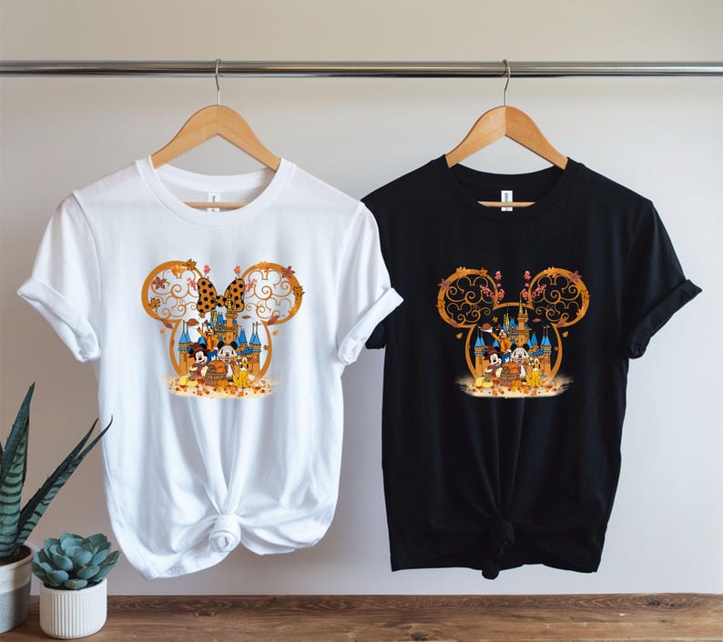 Pode incluir: Duas camisetas, uma branca e outra preta, cada uma com um design tem&aacute;tico da Disney. O design inclui um la&ccedil;o da Minnie Mouse, folhas de outono e personagens da Disney em frente a um castelo. As camisetas est&atilde;o penduradas em cabides de madeira.