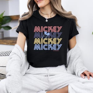 May include: Black t-shirt with the word "MICKEY" stacked four times in pink, purple, yellow, and blue. A silhouette of Mickey Mouse is in the second "MICKEY". The text "THE ORIGINAL" is below the stacked words. The shirt is a casual wear item.