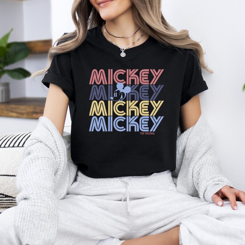 May include: Black t-shirt with the word "MICKEY" stacked four times in pink, purple, yellow, and blue. A silhouette of Mickey Mouse is in the second "MICKEY". The text "THE ORIGINAL" is below the stacked words. The shirt is a casual wear item.