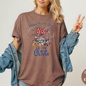 Pode incluir: Camiseta marrom com um gr&aacute;fico com Mickey e Minnie Mouse em trajes patri&oacute;ticos, um la&ccedil;o vermelho e o texto "Happy 4th of July" e "Olivia". A camisa &eacute; uma cor de conforto em espresso.