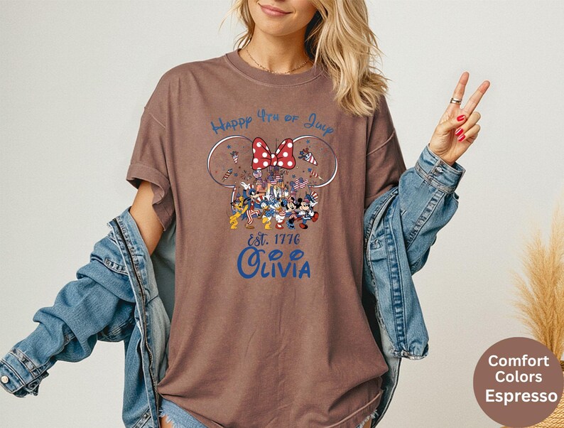 Pode incluir: Camiseta marrom com um gr&aacute;fico com Mickey e Minnie Mouse em trajes patri&oacute;ticos, um la&ccedil;o vermelho e o texto "Happy 4th of July" e "Olivia". A camisa &eacute; uma cor de conforto em espresso.