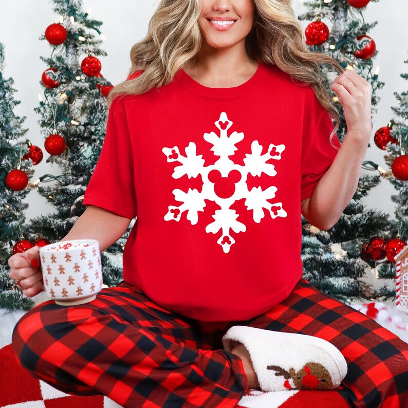May include: A red t-shirt with a white snowflake design featuring a Mickey Mouse head in the center. The person is wearing red and black plaid pajama pants and white slippers. Christmas trees with red ornaments are in the background.