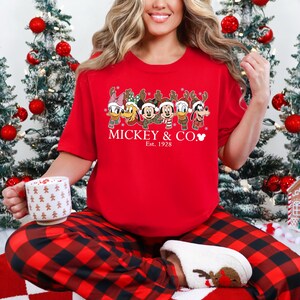 K&ouml;nnte beinhalten: Rotes kurz&auml;rmeliges T-Shirt mit einem festlichen Design von Disney-Figuren mit Rentiergeweih und Schals. Der Text "MICKEY & CO." und "Est. 1928" sind unter den Figuren aufgedruckt. Die Person tr&auml;gt eine rot-schwarz karierte Pyjamahose.