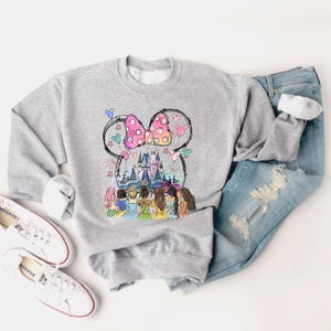May include: A gray sweatshirt with a graphic print of a cartoon mouse head with a bow and the Disney castle in the background. The graphic also includes a group of cartoon princesses in front of the castle.