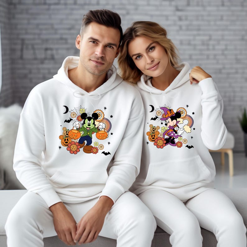K&ouml;nnte beinhalten: Wei&szlig;e Kapuzenpullover mit passenden Jogginghosen. Die Sweatshirts zeigen Halloween-Cartoonfiguren, darunter Micky Maus und Minnie Maus, mit K&uuml;rbissen, Regenbogen und Flederm&auml;usen. Ideal f&uuml;r Halloween.