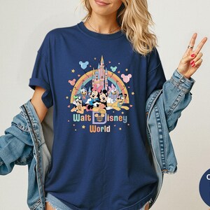 Puede incluir: Camiseta azul marino con un gr&aacute;fico colorido de personajes de Disney, un arco&iacute;ris y un castillo. El texto "Walt Disney World" se muestra debajo de los personajes. La camiseta tiene mangas cortas y cuello redondo.