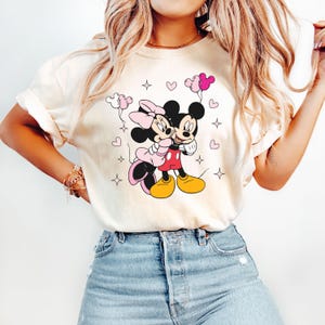 May include: A white t-shirt with a graphic of Mickey Mouse and Minnie Mouse hugging. They are surrounded by pink hearts and stars. Minnie Mouse is wearing a pink bow and Mickey Mouse is wearing red shorts.