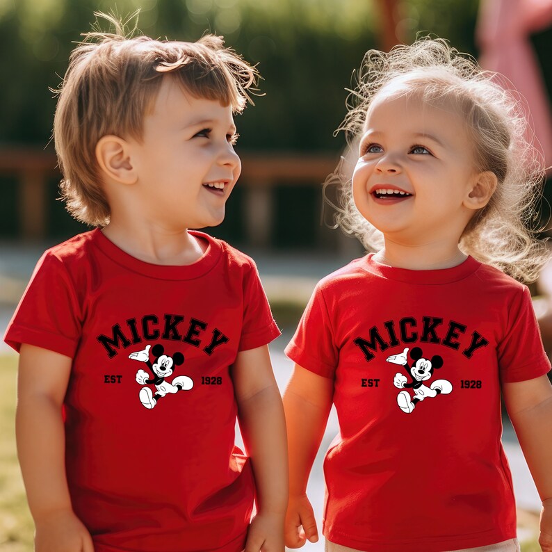 May include: Two children wearing red t-shirts with a black Mickey Mouse graphic and the text "Mickey Est 1928".