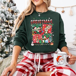 May include: A dark green sweatshirt with a festive Christmas design featuring cartoon characters. The words "Very Merry Christmas" are displayed above the graphic. The person is wearing red and white plaid pajama pants and holding a mug with a Santa Claus design.